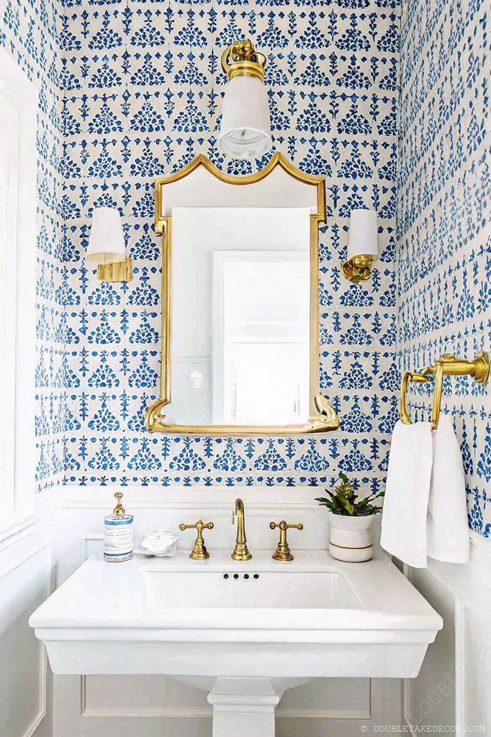 Salle de bain avec crédence à fleurs bleues et blanches, miroir à contours ondulés, robinetterie dorée et accessoires assortis pour une ambiance sophistiquée.