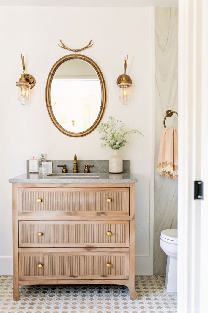 Meuble-vasque en bois clair avec plan de travail gris, miroir ovale doré et appliques assorties pour une salle de bain élégante et moderne.