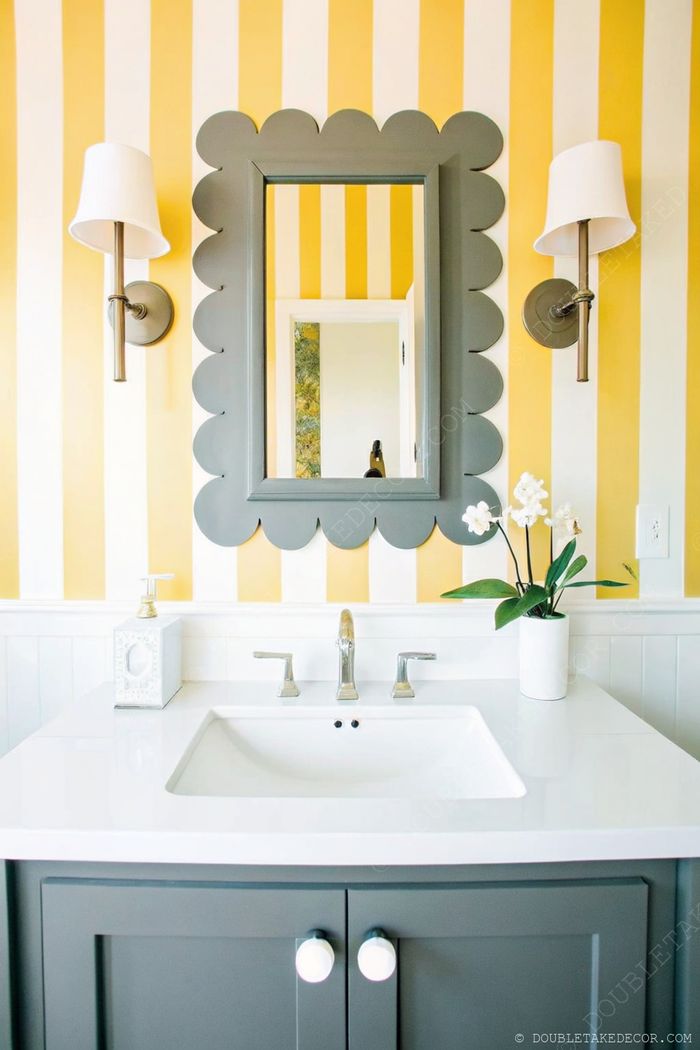 Petite salle d’eau moderne avec murs à rayures jaunes et blanches, miroir à bords ondulés, meuble gris et touches de plantes.