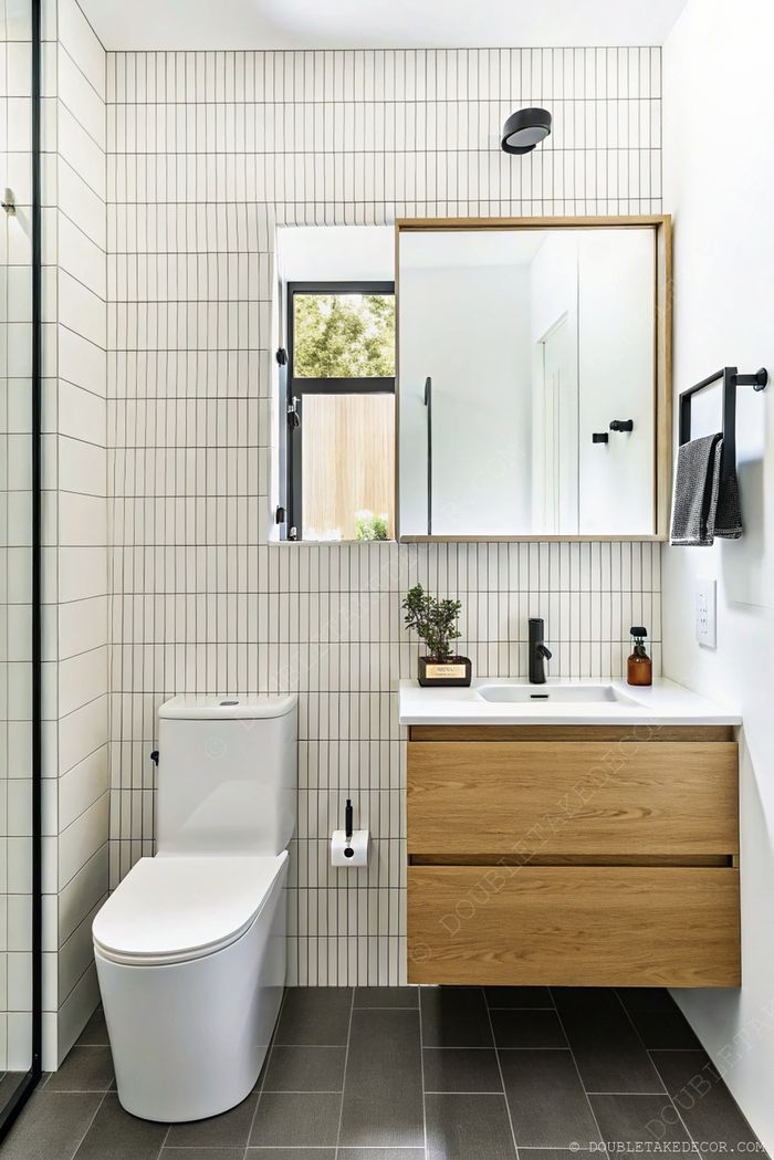 Salle de bain minimaliste avec carreaux clairs, douche vitrée, toilettes blanches, meuble-vasque en bois, robinet noir et lumière naturelle.