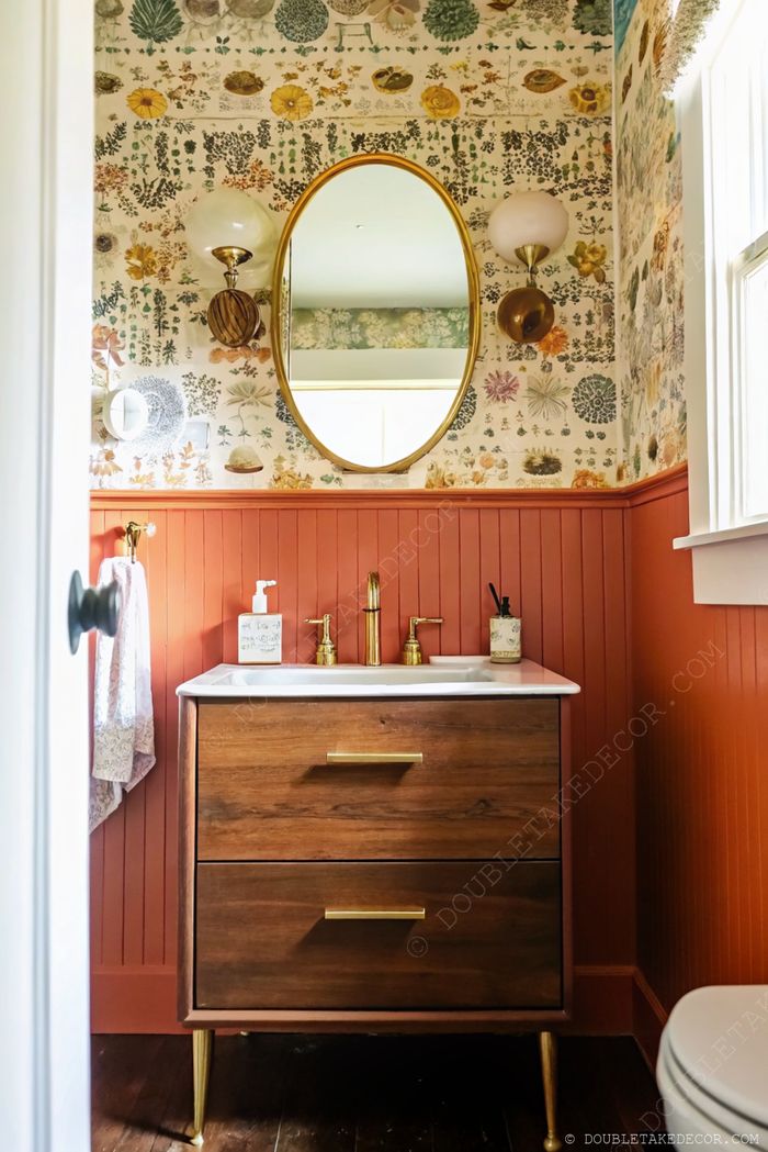 Petite salle de bain stylée avec papier peint floral coloré, miroir doré, meuble en bois et soubassement orange-rouge.