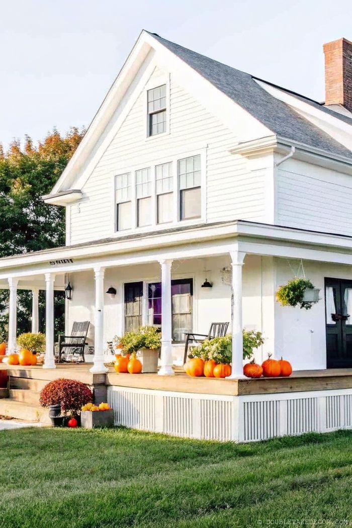 White house with slender columns featuring seasonal orange pumpkins, black rocking chairs and potted plants on a wraparound porch.