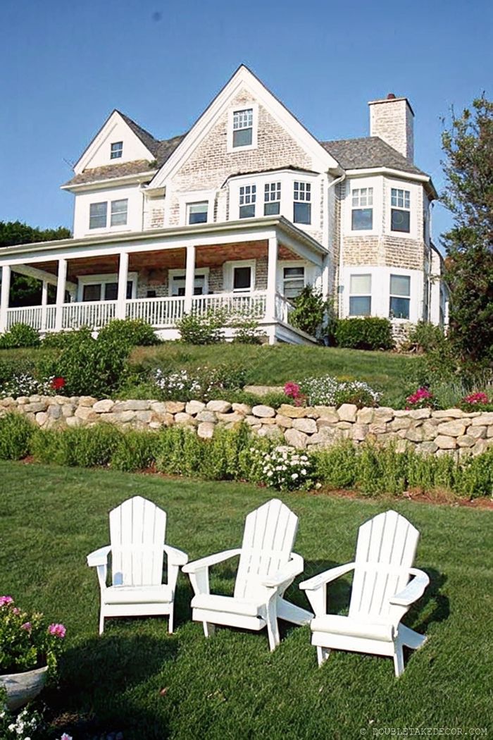 Multi-story home with wraparound porch and white Adirondack chairs on lawn surrounded by colorful flower beds