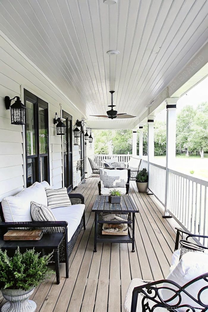Inviting wraparound porch with wooden decking, white railings, wicker sofas with plush cushions, and hanging black lanterns.