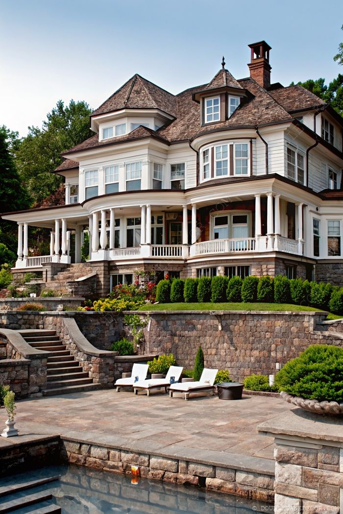 Elegant wraparound porch home with stone and wood exterior, featuring columns, tiered landscaping and a serene pool area with lounge chairs.