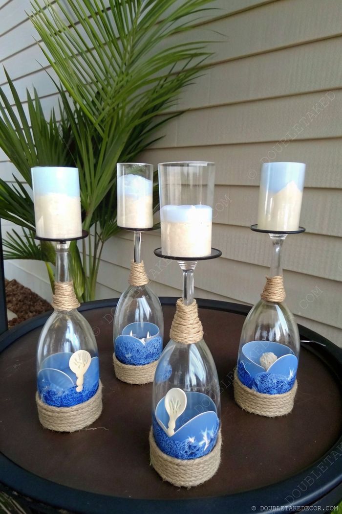 Decorative candle holders made from inverted wine glasses adorned with twine and filled with blue beach-inspired layers, creating coastal charm.