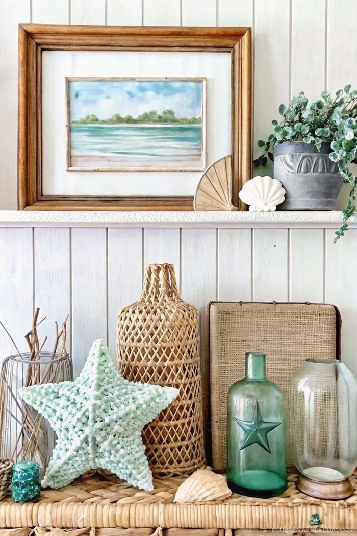 Coastal themed shelf with a watercolor painting, seashells, a mint green star, woven baskets, and vintage glass bottles arranged on a light woven mat.