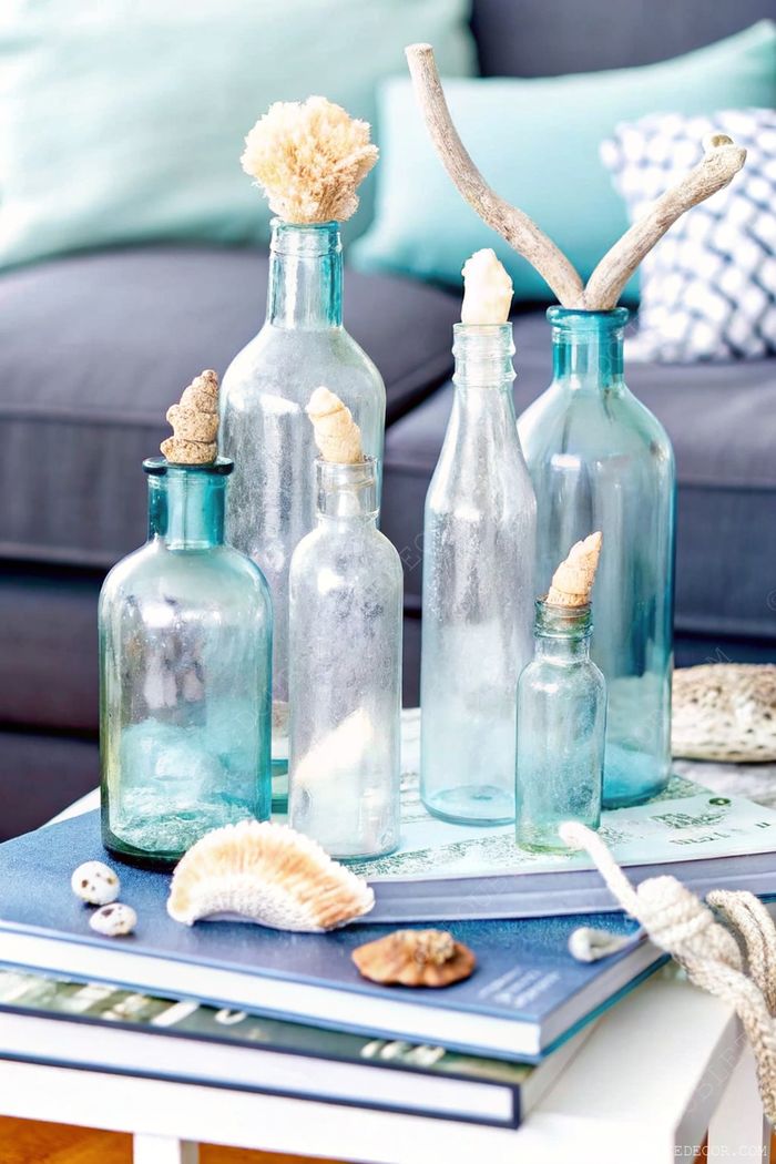 Coastal-inspired decor featuring blue glass bottles with dried flowers and coral, arranged with seashells on a gray couch with turquoise pillows.