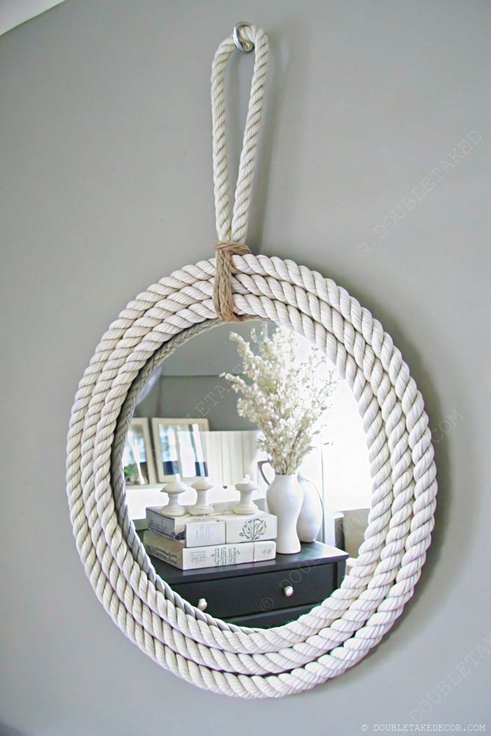 Round mirror with white rope frame hung above a dark chest adorned with books, candlesticks, and dried flowers in a vase