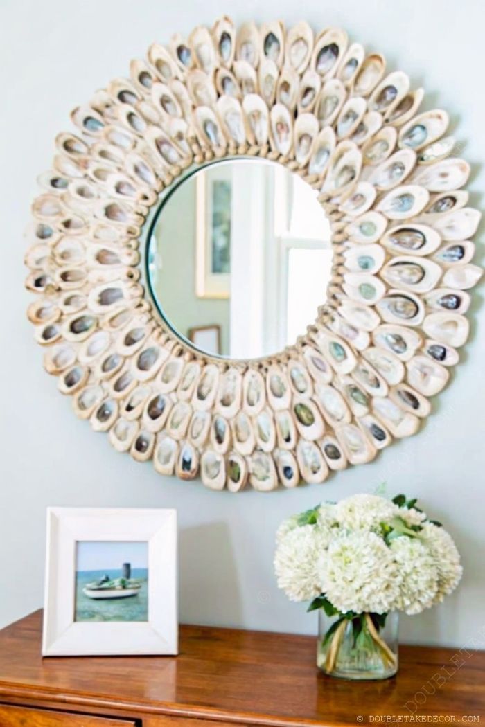 A round mirror with shell-inspired frame above a wooden console displaying a boat photograph and white flower arrangement in a coastal color palette.