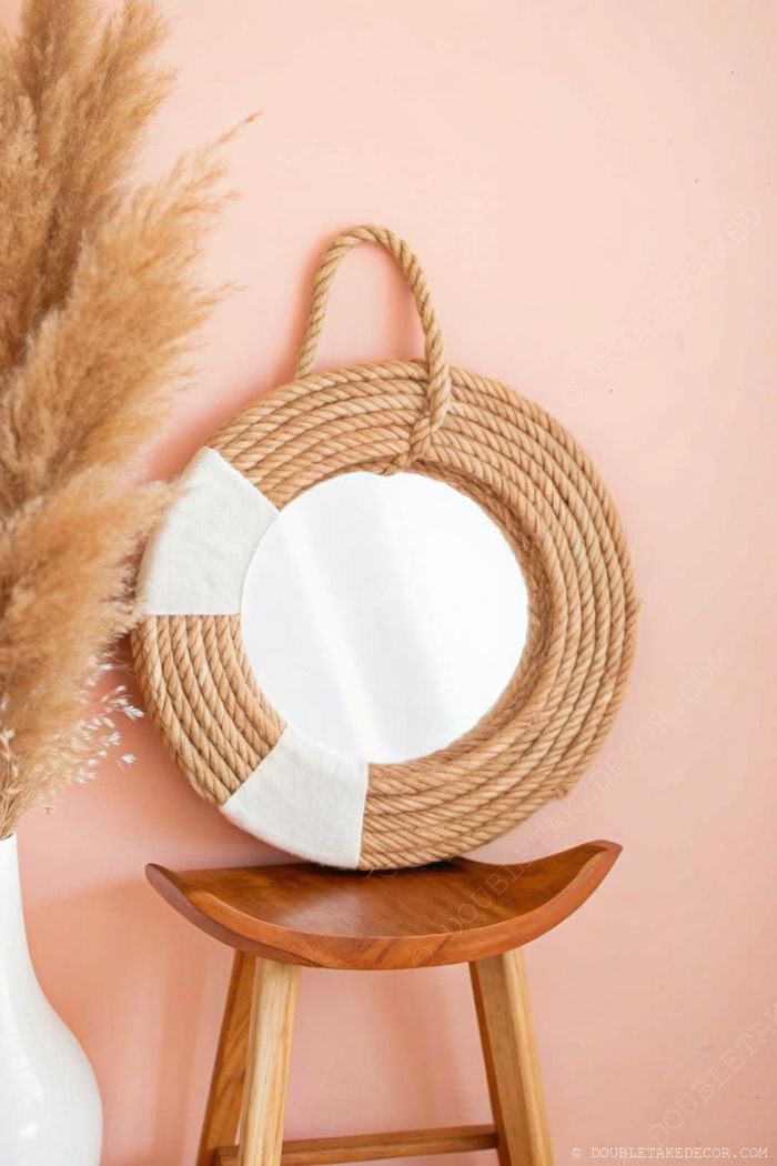 Round mirror with natural jute rope frame and white fabric accents against peach wall with pampas grass vase on wooden stool