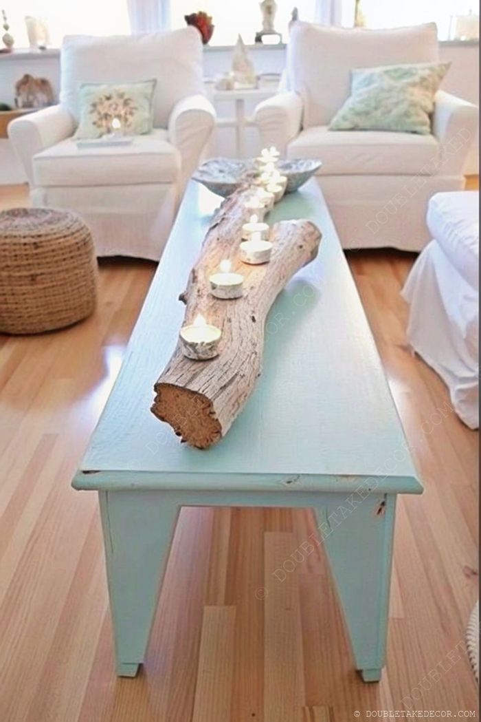 A coastal-inspired living room featuring a pale blue coffee table with a driftwood log centerpiece adorned with white candles, surrounded by white armchairs.