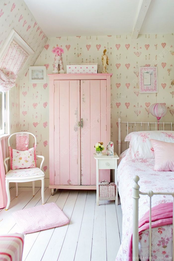 A charming bedroom with soft pink walls, distressed wooden wardrobe, floral bedding, and a cozy white armchair creating the perfect shabby chic sanctuary.