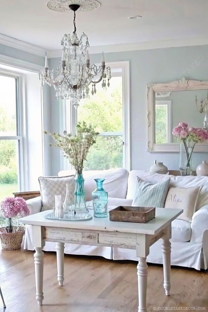 Levegős nappali kopottas fehér dohányzóasztallal, puha kanapéval, pasztell dekorral, friss virágokkal és elegáns csillárral, shabby chic stílusban.