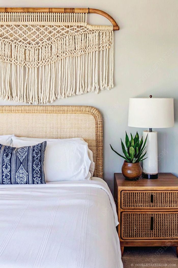 Bedroom with natural woven headboard, macramé wall art, white linens, and rattan nightstand holding a lamp and plant.