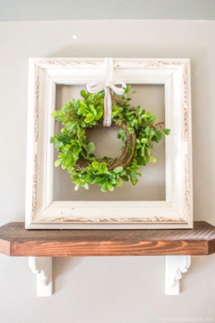 Green wreath in white vintage frame with ribbon, displayed on wooden shelf with white brackets against gray wall