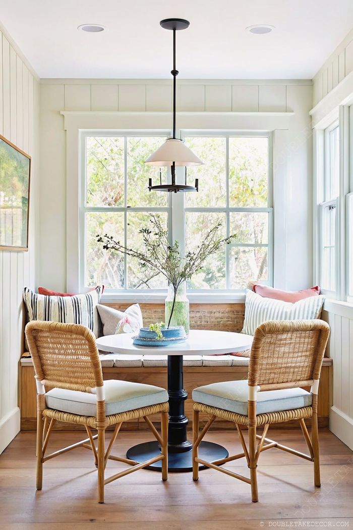 Cozy breakfast nook with round white table, rattan chairs, built-in bench with colorful pillows, and natural light.