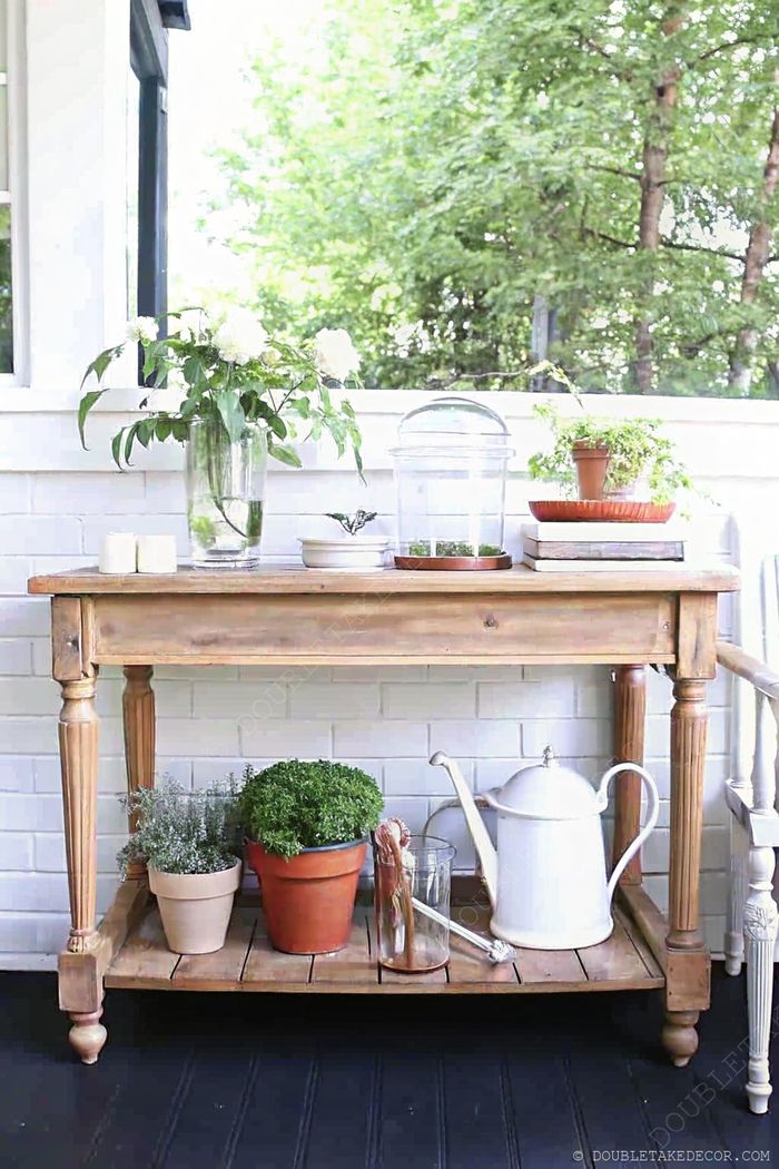 Rustic outdoor table with white flowers, potted herbs, and gardening accessories against a white brick wall.