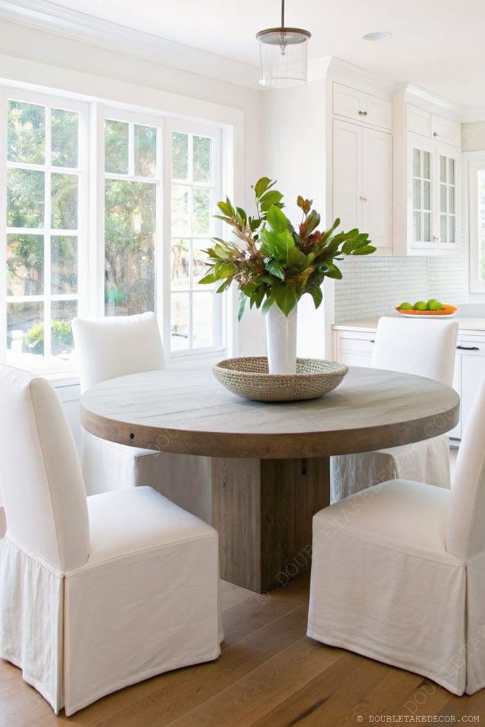 Rustic round wooden table with white slipcover chairs, greenery centerpiece, and natural light creating a warm farmhouse dining area