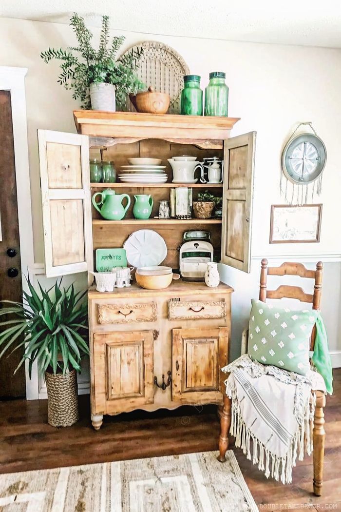 Cozy farmhouse interior with wooden hutch displaying green glassware, white plates, and rustic decorative elements