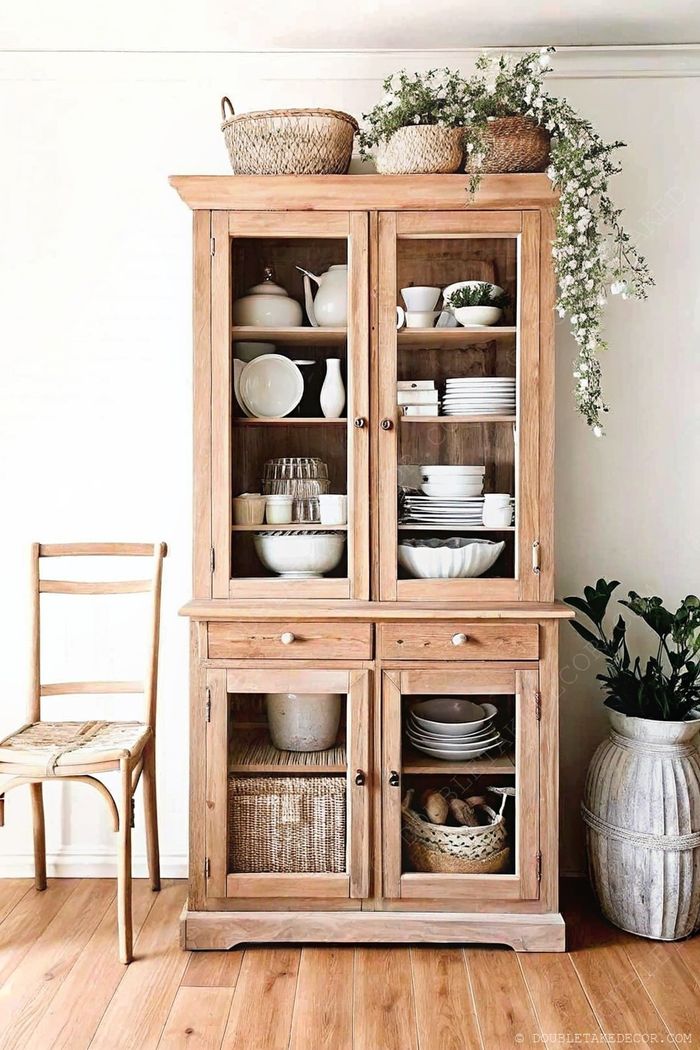 Wooden cabinet with glass doors displaying organized dishes, topped with baskets and greenery, complemented by a woven chair