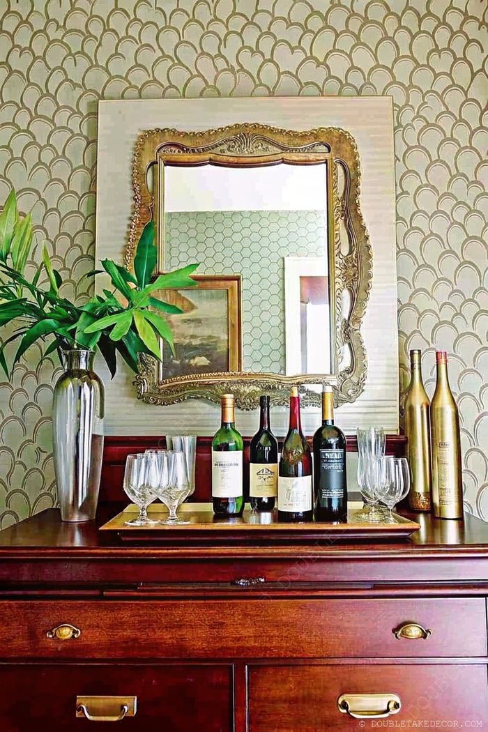 Wooden sideboard styled as a bar with wine bottles, glassware, and plant against scalloped farmhouse wallpaper with decorative mirror.