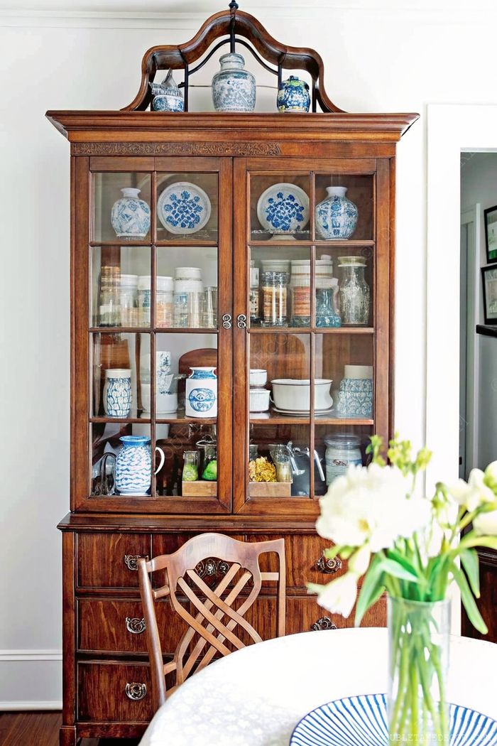 Elegant wooden hutch with glass doors displaying ceramic jars, blue floral plates, and topped with decorative pieces beside a dining table with white flowers.