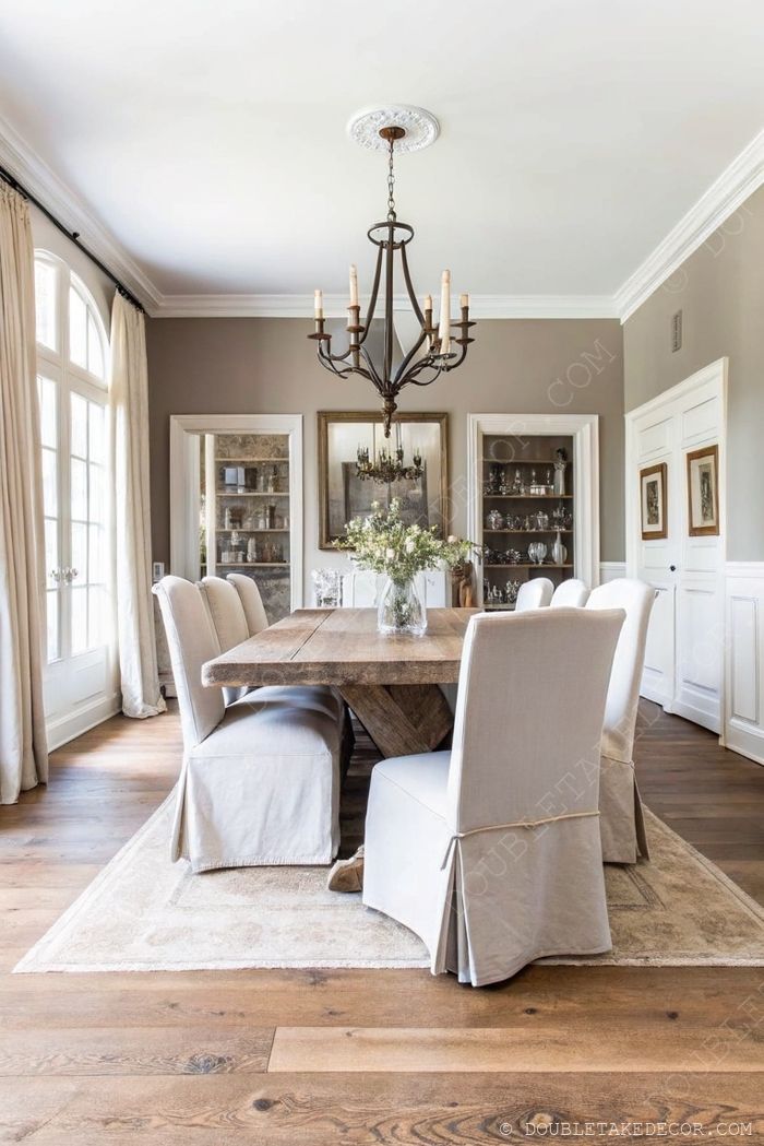Wooden farmhouse table with upholstered chairs under candle chandelier in neutral dining room with china cabinets