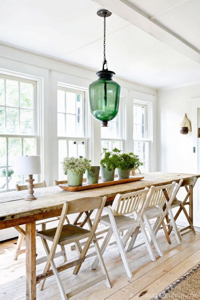 Airy dining space with rustic wooden farmhouse table, white chairs, potted plants, and striking green pendant light.