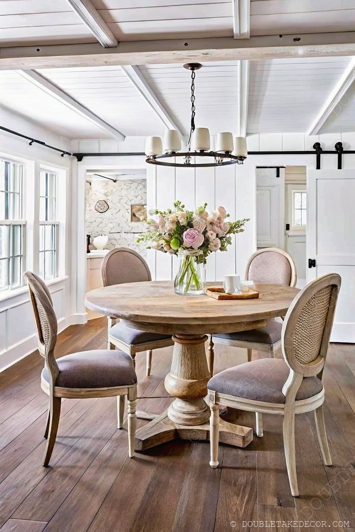Elegant round wooden pedestal table with upholstered chairs, chandelier above, and floral centerpiece in a bright dining space.