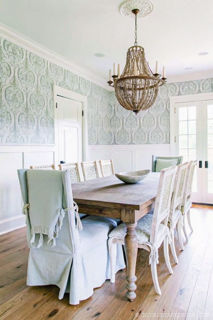 Rustic wooden farmhouse table surrounded by blue fringed chairs, beneath a wicker chandelier with green wallpaper backdrop.