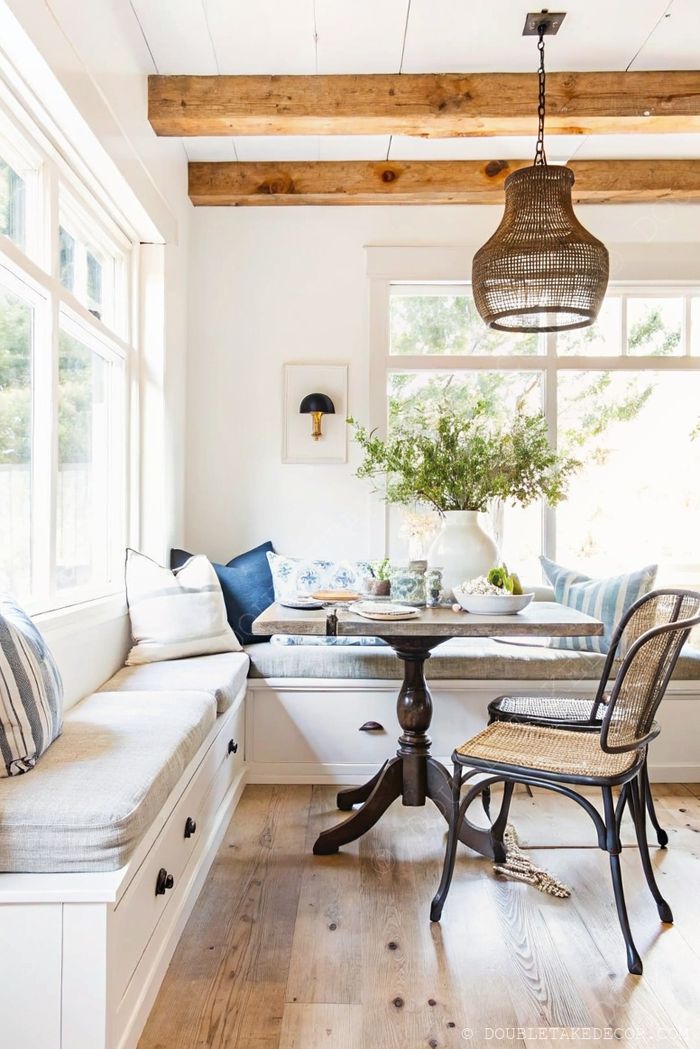 Bright farmhouse dining nook with cushioned bench seating, wooden pedestal table, woven pendant light, and natural wood beams.