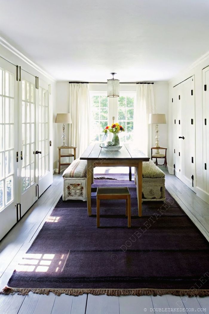 Rustic wooden farmhouse table in a bright dining area with sheer curtains, textured bench seating, and purple area rug