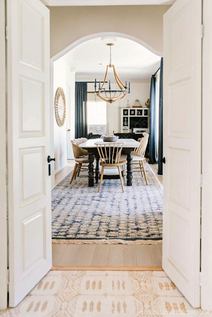 White farmhouse table with mixed chairs viewed through archway, featuring gold chandelier, patterned rugs, and dark curtains