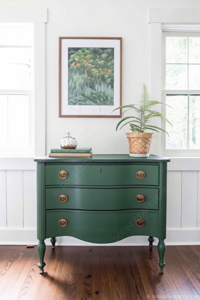Elegante grüne Vintage-Kommode mit Messingknöpfen, dekoriert mit Büchern, Farn im Topf und Naturbildern in einem hellen, luftigen Raum.