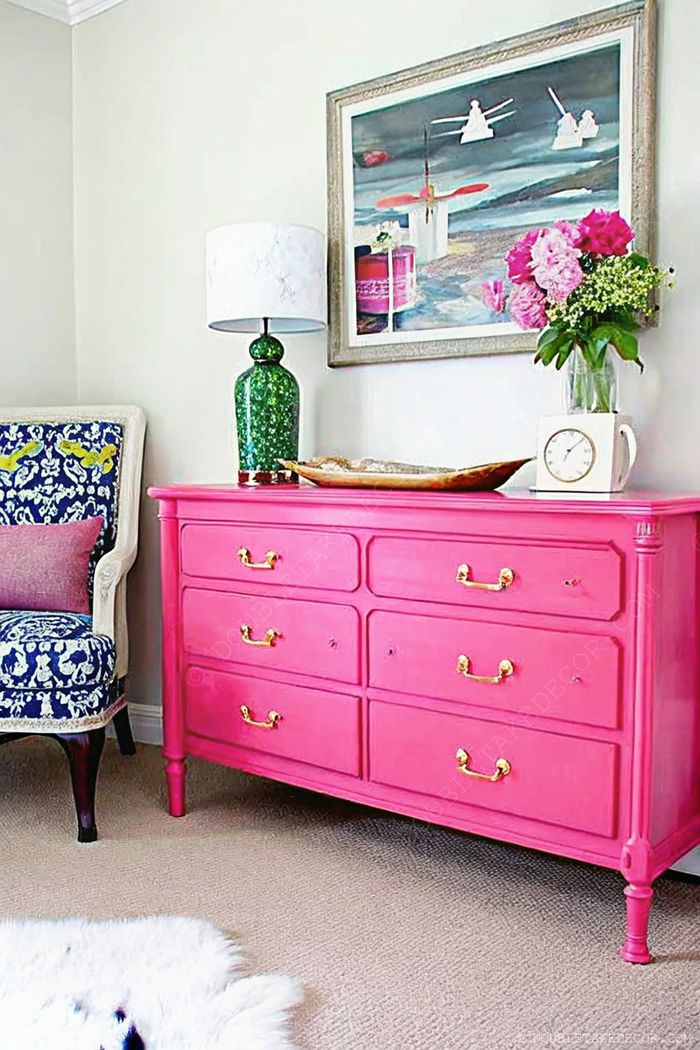 Farbenfrohes Interior mit knallpinker Kommode und goldenen Griffen, dekorativen Accessoires und blau gemustertem Sessel für fröhliche Stimmung.