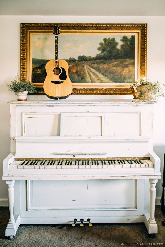 Weißes, abgenutztes Klavier mit Landschaftsbild darüber, Gitarre an der Seite und kleinen Pflanzen als rustikales Arrangement.