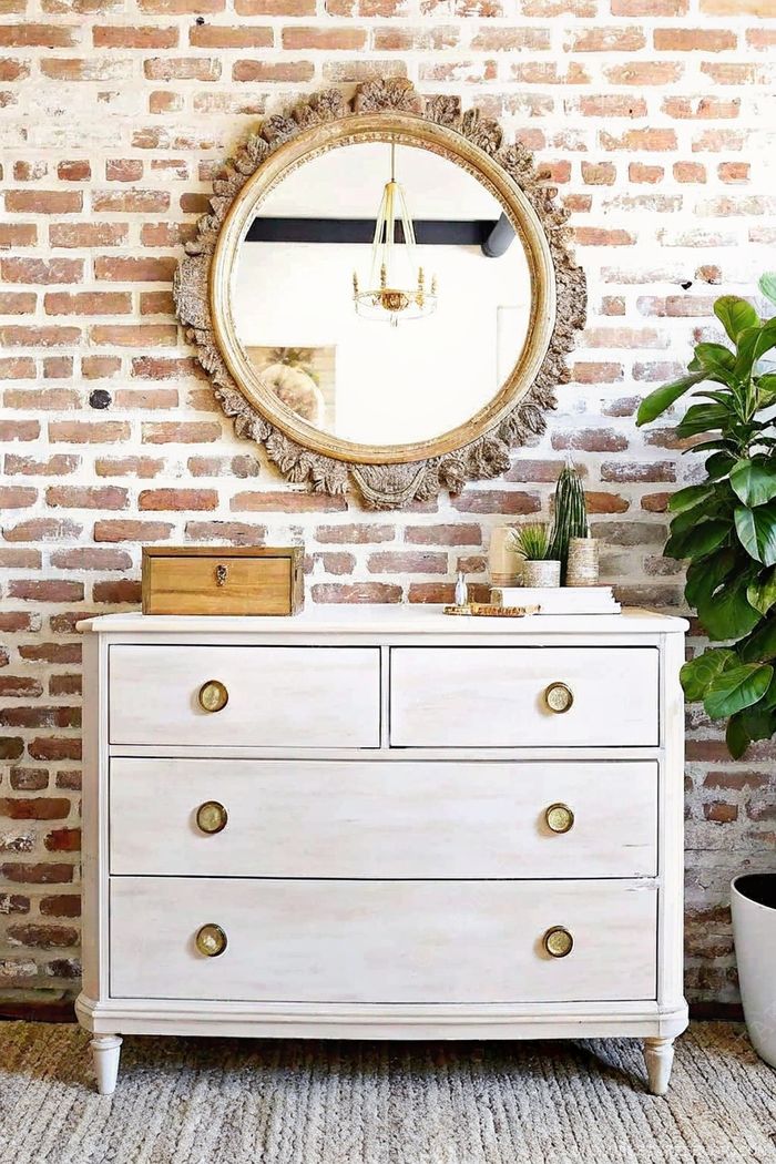 Weiße Kommode mit goldenen Knöpfen vor Backsteinwand, dekoriert mit Zubehör und rundem Spiegel.