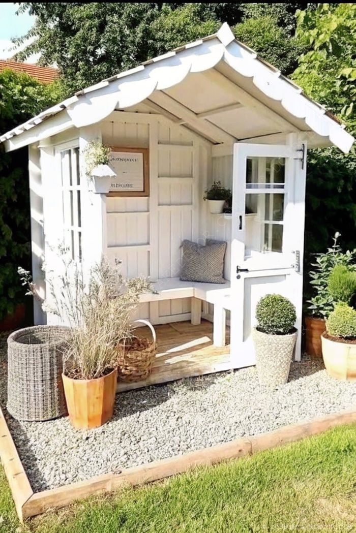 Un joli abri de jardin blanc avec portes vitrées, banquette à l'intérieur et pots de fleurs tout autour – le coin parfait pour se détendre.