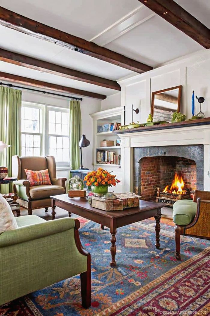 Warm living room with wooden beams, featuring green curtains, comfortable armchairs, and a fireplace with brick surround.
