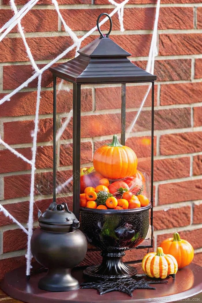 Metal lantern filled with pumpkins and mandarins, displayed with a black cauldron against brick backdrop with spider web accents.