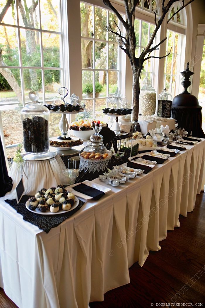 Sophisticated Halloween dessert table with dark treats on black lace, glass jars, and spooky tree branch against white tablecloth and window backdrop.