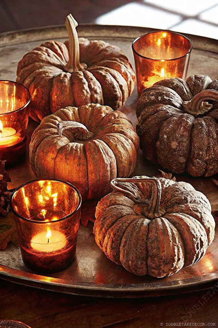Rustic silver tray displaying various textured pumpkins surrounded by glowing amber glass candles, creating warm autumn ambiance.