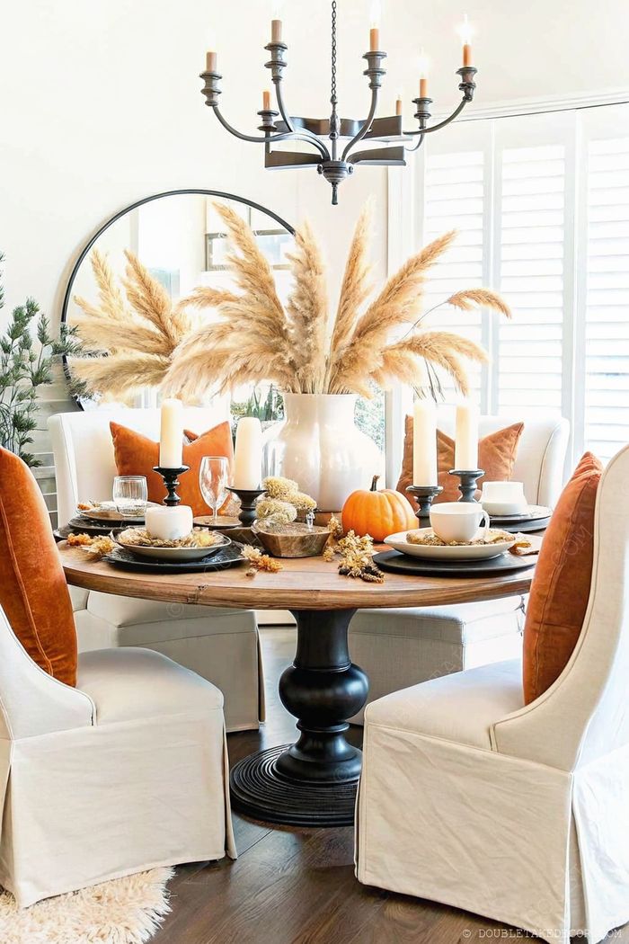 Round wooden table decorated with pampas grass, black candle holders, and seasonal elements like pumpkins for a sophisticated Halloween dining experience.