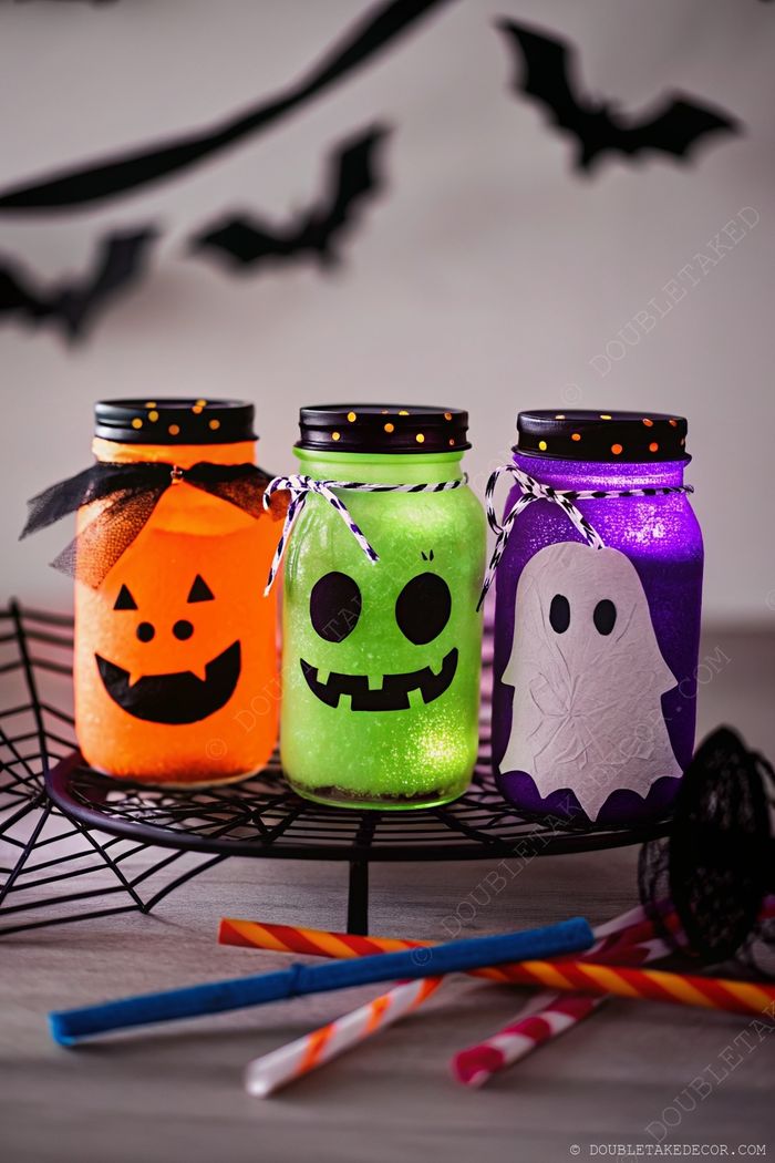 Illuminated Halloween jars designed as a pumpkin, monster, and ghost, arranged on a spiderweb stand with colorful straws for festive table decor.