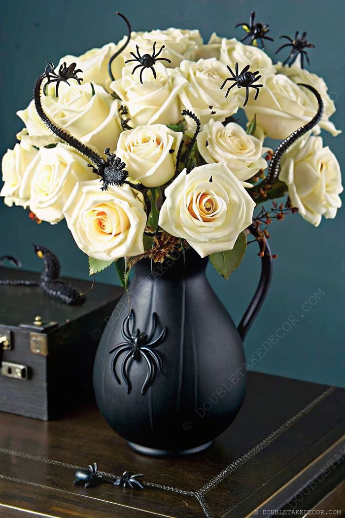 Elegant white roses in a black vase with spider motif, surrounded by decorative black spiders for a sophisticated Halloween table decoration.