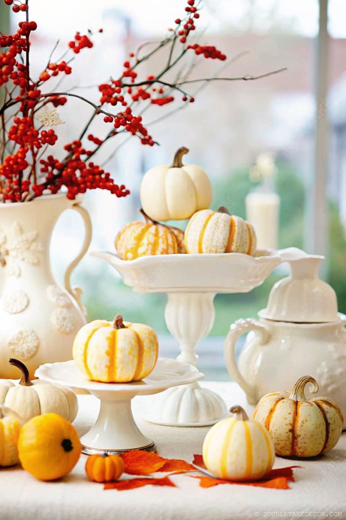 A charming table display with decorative white and striped pumpkins on cake stands, accompanied by a white pitcher with red berry branches