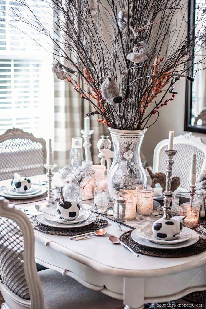 Elegant Halloween dining table with a centerpiece of bare branches, bird decorations, and candles, featuring cow-patterned bowls on white plates.
