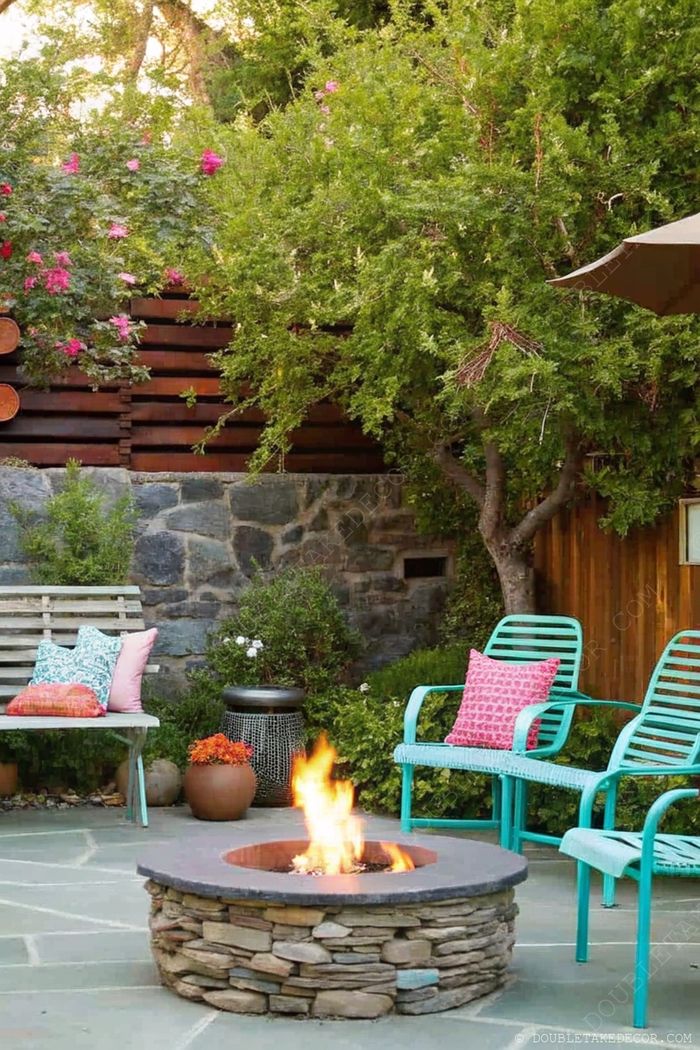 Cozy outdoor fire pit with natural stone border, turquoise chairs with accent pillows, and wooden bench surrounded by lush greenery.