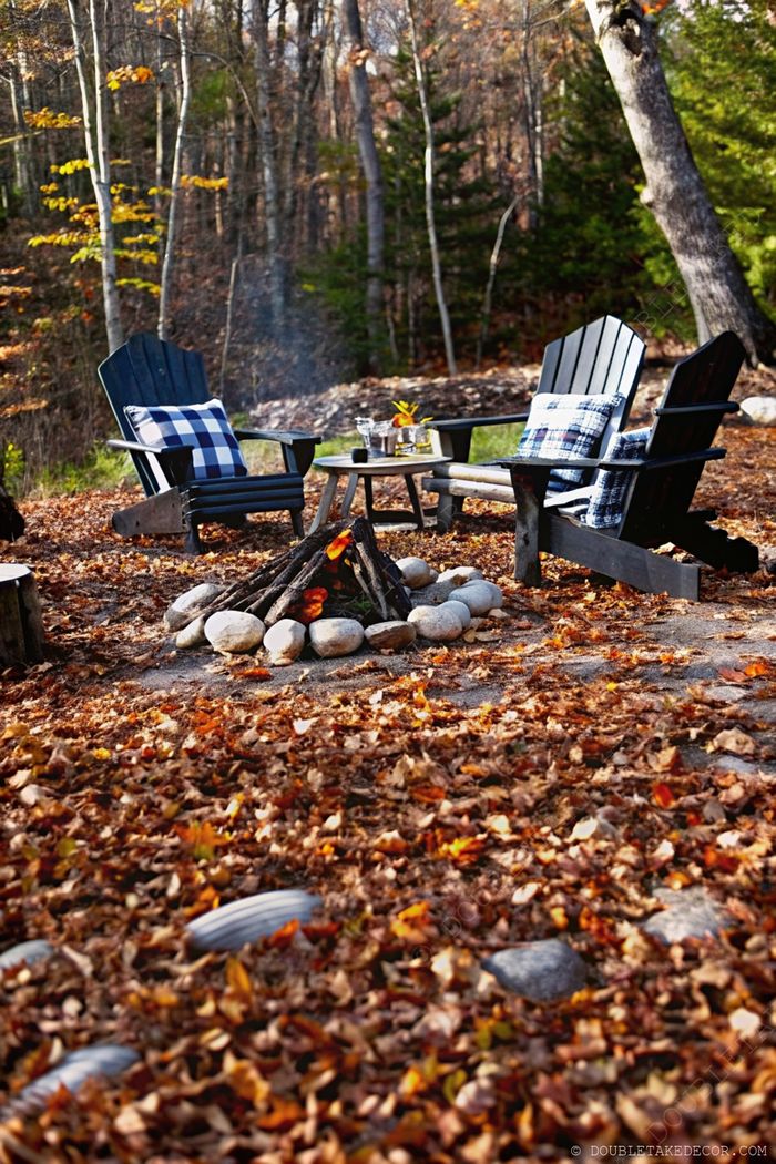 Cozy black Adirondack chairs with blue checked pillows beside a stone-encircled fire pit, surrounded by colorful fallen leaves and autumn foliage.